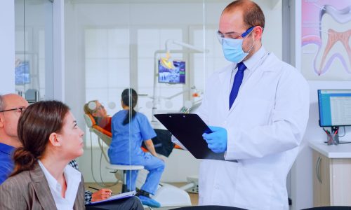 Doctor taking notes on clipboard about patient dental problems sitting on chair in waiting room of stomatological clinic. Assistant preparing old woman for examination in background